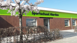 De voorkant van het gebouw van Sportschool Hierden (Witte bovenkant met een groene strook met zwarte letters 'Sportschool Hierden', daaronder een houten muurplaat. Voor de sportschool staat een heg met een Japanse bloessem boom.
